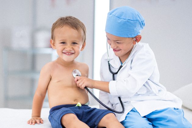 An adorable Eurasian toddler boy smiles directly at the camera as his older brother, who is dressed up in a doctor costume, uses a stethoscope to check his heart and lungs.