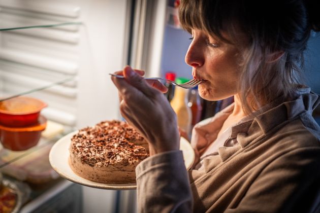 Mature woman eating cake while standing near open refrigerator at home.