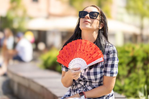 Woman in sunglasses faces sun and uses spread or hand fan in the summer heat in the city.