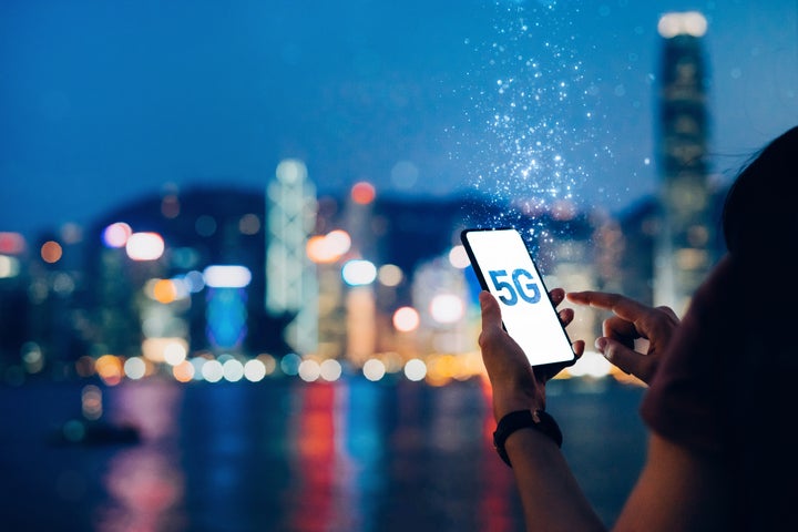 Junge Frau nutzt ein Smartphone mit der Aufschrift „5G“ vor der berühmten Skyline von Hongkong neben der Victoria Harbour Promenade bei Nacht, wobei das Konzept der 5G-Kommunikation in Lichtpartikeln aufgelöst ist