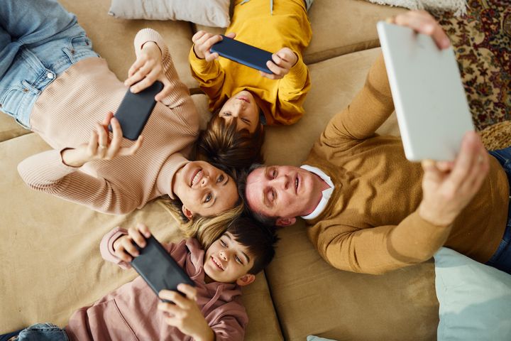 Blick aus der Vogelperspektive auf eine glückliche Familie, die drahtlose Technologie nutzt, während sie zu Hause auf dem Sofa liegt