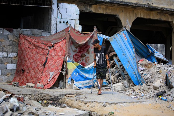 2024 年 6 月 30 日，一名受伤的巴勒斯坦男子拄着拐杖走在收容难民的帐篷附近，该帐篷搭建在先前以色列袭击中被毁的建筑物前，一些人正在返回加沙南部城市汗尤尼斯。