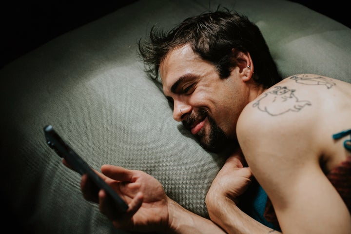 A tattooed man giggles and blushes as he coyly looks at his smart phone as he lies on his side in bed.