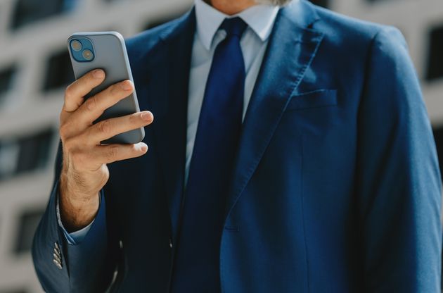 Close-up of a mature businessman in a blue suit using a smartphone while standing outdoors in a modern urban setting.