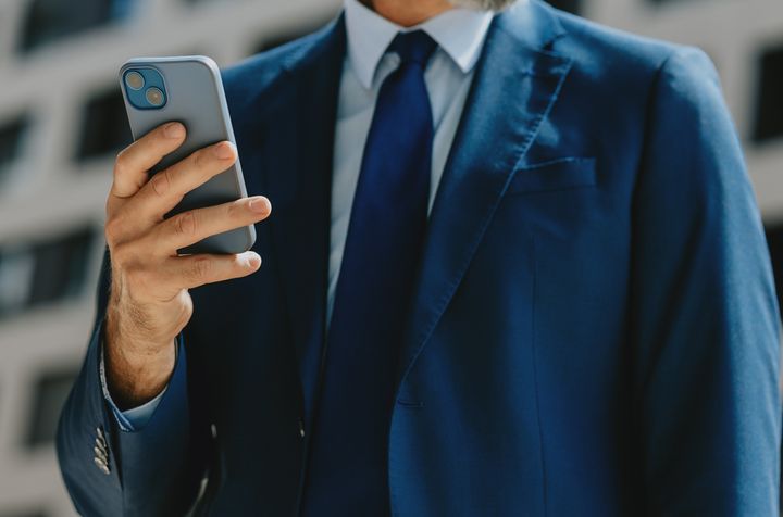 Close-up of a mature businessman in a blue suit using a smartphone while standing outdoors in a modern urban setting.