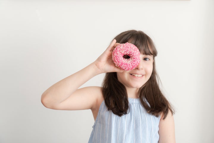 Little girl is eating pink doughnut with sprinkles in front of white background.