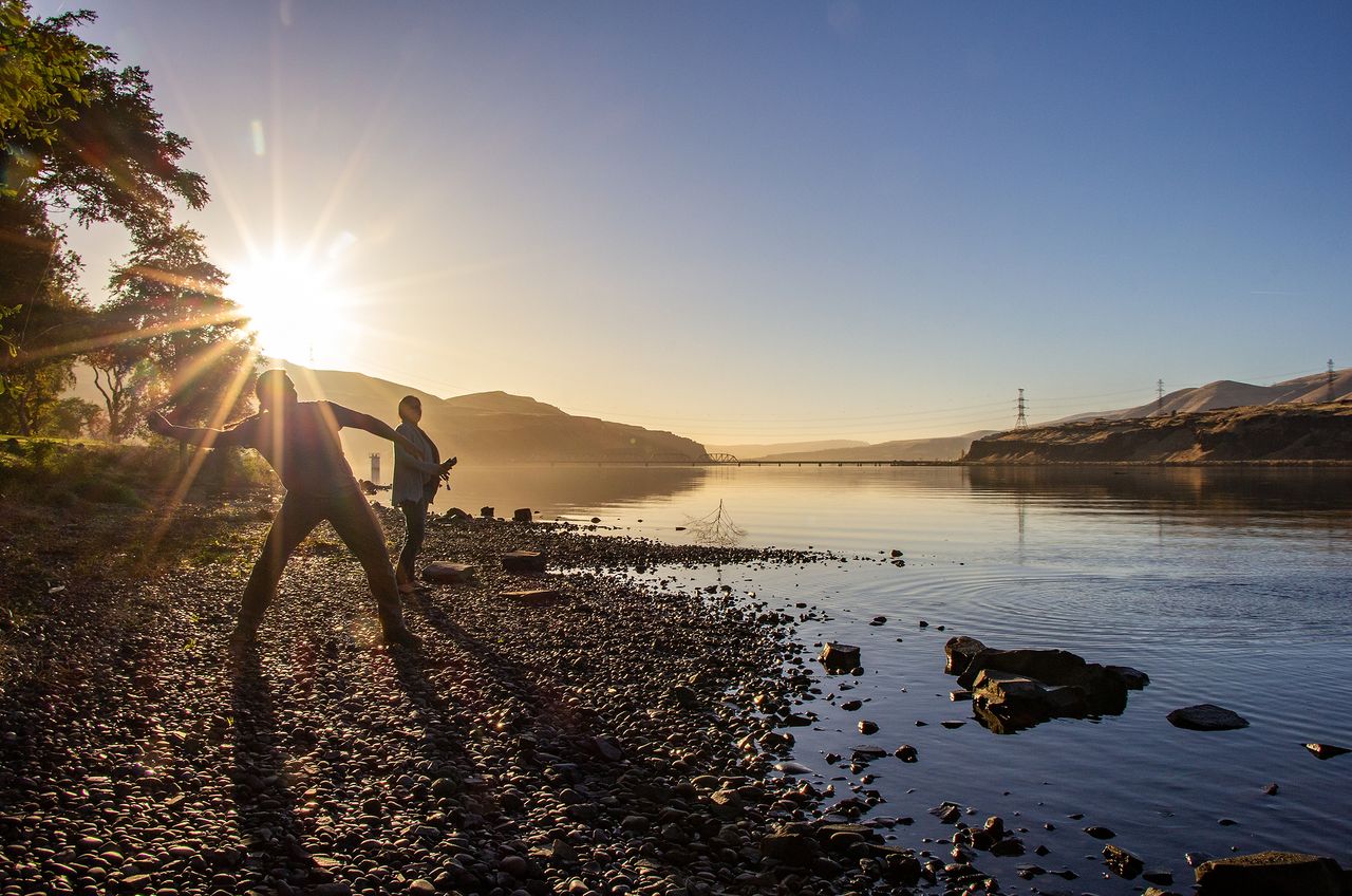 Andrew Wildbill (Hinmat Kakykt - Thunder Strikes) Cayuse, and Ashley Harding, Navajo, skip rocks on the Columbia River at Celilo Park, Oregon. The slackwater shown here sits behind The Dalles Dam, which displaced an entire Indigenous village and devastated the Columbia River ecology. Andrew is the Program Manager for the Confederated Tribes of the Umatilla Indian Reservation Natural Resources Department Wildlife Program. Their tribal government has been a key player in the protection and recovery of the Columbia River watershed by using treaty rights to force compliance with fish passageway requirements in the Columbia River dams, and other protections for the region's fish and wildlife. They have implemented countless programs to help recover threatened and endangered species in the Pacific Northwest. They are guided by principles of sustainable and reciprocal relationship with nature they call Tamanwit.