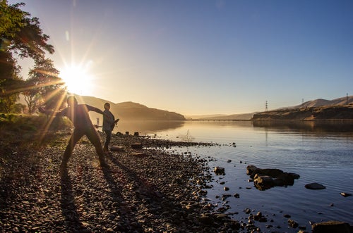 Andrew Wildbill (Hinmat Kakykt - Thunder Strikes) Cayuse, and Ashley Harding, Navajo, skip rocks on the Columbia River at Celilo Park, Oregon. The slackwater shown here sits behind The Dalles Dam, which displaced an entire Indigenous village and devastated the Columbia River ecology. Andrew is the Program Manager for the Confederated Tribes of the Umatilla Indian Reservation Natural Resources Department Wildlife Program. Their tribal government has been a key player in the protection and recovery of the Columbia River watershed by using treaty rights to force compliance with fish passageway requirements in the Columbia River dams, and other protections for the region's fish and wildlife. They have implemented countless programs to help recover threatened and endangered species in the Pacific Northwest. They are guided by principles of sustainable and reciprocal relationship with nature they call Tamanwit.
