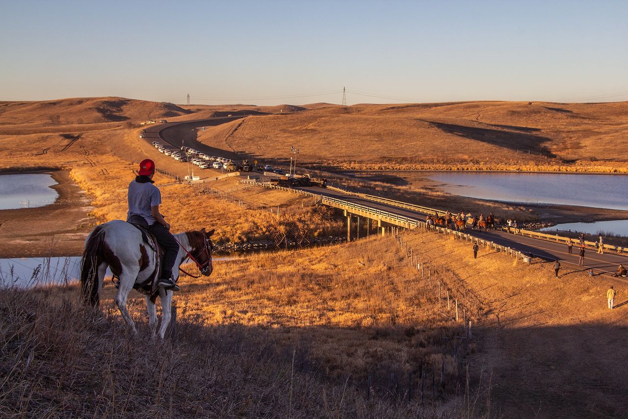 Indigenous “horse warriors” gather on the Backwater Bridge at the Standing Rock Sioux Reservation for a ceremony held during the Dakota Access Pipeline protest movement of 2016. Indigenous activists known as “water protectors” fought back for months against a tar sands pipeline that was forced through the reservation’s drinking water. Two weeks after this photo was taken, the paramilitary police and mercenaries depicted here blockading the other side of the bridge, sprayed water cannons on hundreds of Indigenous People gathered in prayer on the bridge in below freezing temperatures, giving over 140 people hypothermia, and partially blinding one woman and tearing the lower arm off of another woman with “nonlethal” armaments. Since its completion the Dakota Access Pipeline has ruptured and leaked multiple times, just as water protectors predicted it would.