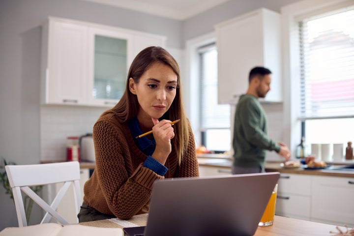 Mid adult woman using laptop while working at home. Her husband is in the background.