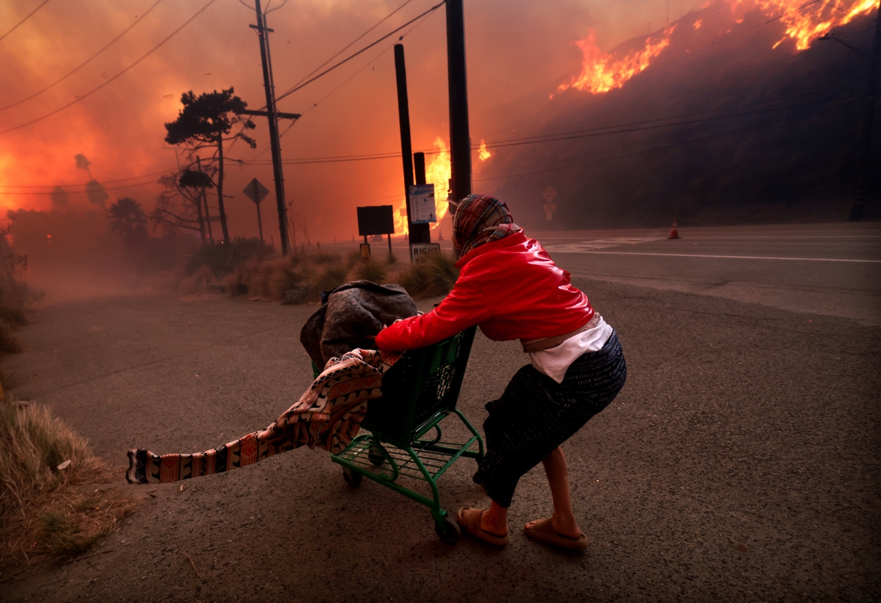 A homeless woman pushes her belongings off the Pacific Coast Highway and Topanga Canyon Boulevard as the Palisades Fire rages down the hills in the Pacific Palisades on Tuesday.