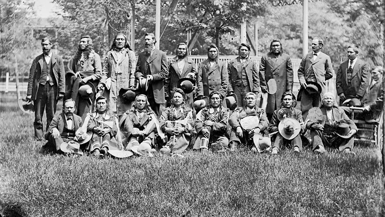 Thirteen Native American men, probably visiting chiefs, pose for a portrait with five white men and Richard Henry Pratt (seated in chair at right).