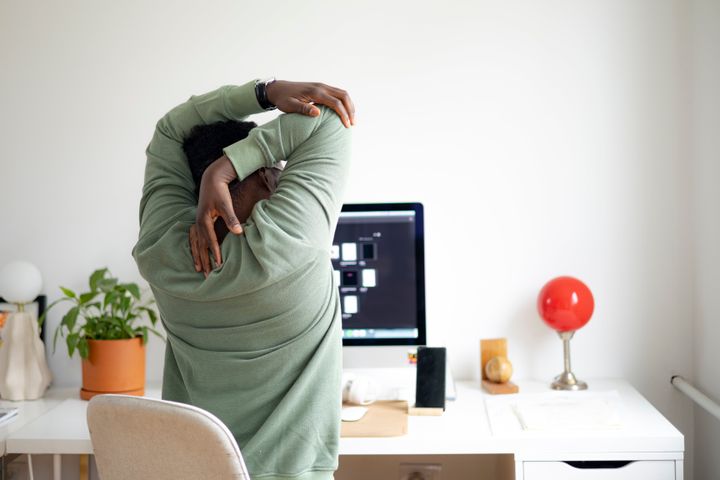 Rear view of a male freelancer having back problems while sitting at his desk in front of the monitor, feeling the pain in his back and shoulders