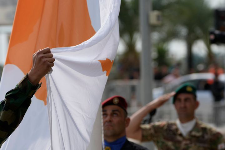 FILE - A police officer holds a Cyprus' flag as two soldiers salute during a ceremony before a military parade marking the 63th anniversary of Cyprus' independence from British colonial rule, in divided capital Nicosia, Cyprus, on Sunday, Oct. 1, 2023. (AP Photo/Petros Karadjias, File)