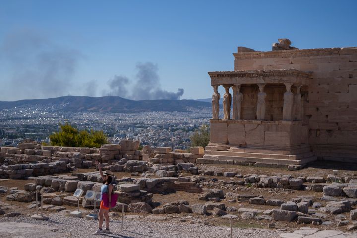 ΑΡΧΕΙΟ - Μια γυναίκα ποζάρει για μια φωτογραφία μπροστά από τα αγάλματα των Καρυάτιδων που στηρίζουν τoν ναό του 5ου αιώνα π.Χ. του Ερεχθείου, καθώς στο βάθος φαίνεται καπνός από φωτιά , κατά τη διάρκεια μιας ζεστής, θυελλώδους ημέρας στο λόφο της Ακρόπολης, στην Αθήνα, Τρίτη 16 Ιουλίου 2024. (AP Photo/Petros Giannakouris, File)