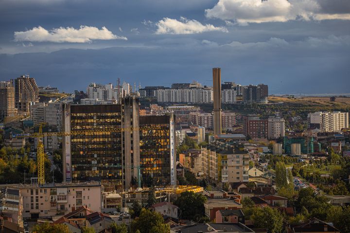 Πρίστινα 2024: Το αστικό τοπίο της Πρίστινα, κάτω από δραματικό συννεφιασμένο ουρανό, με μοντέρνα αρχιτεκτονική και ποικίλα κτίρια.