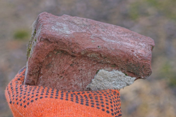 a hand in an orange work mitten holds a piece of red old brick on the street