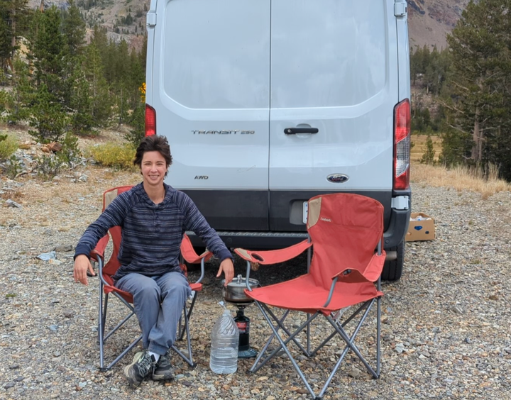 The author relaxing in a camp chair behind the van, with short hair and wearing masculine clothes, in September 2024.