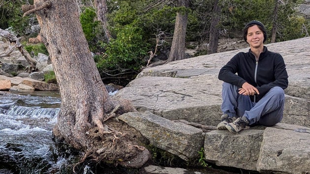 The author sitting beside a mountain stream in August 2024, wearing the same hat, jacket, pants and shoes they had on during the bathroom incident earlier that day.