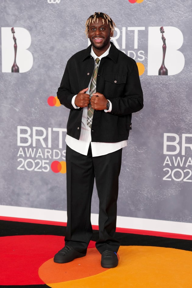 Myles Smith at 2025 Brit Awards: wearing black chunky trainers, slightly schoolboy-chic black trousers, a white shirt, a tie, and a casual jacket