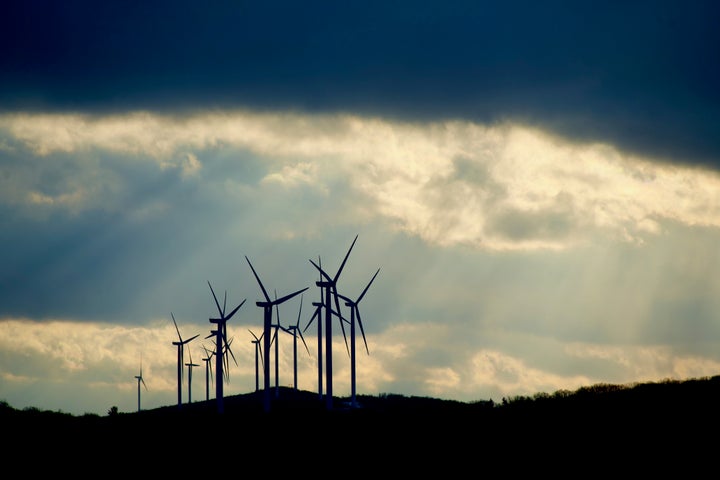 A group of spinning wind turbines line a mountain ridge under storm clouds pierced by afternoon sun.