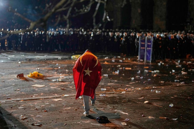 Ένας άνδρας με την τουρκική σημαία στην πλάτη του στέκεται μπροστά από αστυνομικούς των ΜΑΤ κατά τη διάρκεια συγκρούσεων σε διαδήλωση κατά της σύλληψης του δημάρχου της Κωνσταντινούπολης Εκρέμ Ιμάμογλου, στην Κωνσταντινούπολη, Τουρκία, Παρασκευή 21 Μαρτίου 2025. (AP Photo/Khalil Hamra)