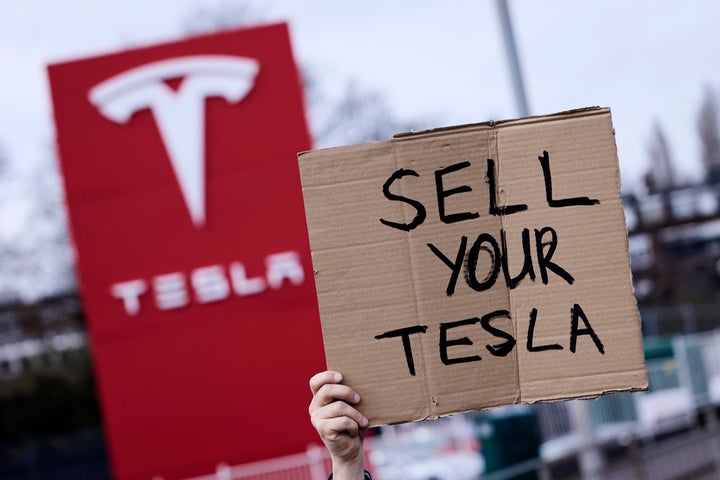 A homemade sign is held up at a protest outside a Tesla dealership.