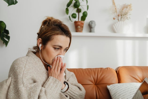 Woman wearing white jumper sitting on terracotta sofa, blowing her nose.