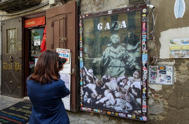 NAPLES, CAMPANIA, ITALY - 2024/03/13: A person takes a picture of British street artist Banksy's 'Madonna with a gun' in the square Gerolamini in the city centre. Unknown perpetrators placed a large stencil with the Pro Palestinian 'Gaza' and a sticker of dead or dying naked children on the glass protecting Banksy's work, without affecting the work, which, when it was discovered in 2016 and found to be original, was covered with a large metal frame to protect it. (Photo by Salvatore Laporta/KONTROLAB/LightRocket via Getty Images)