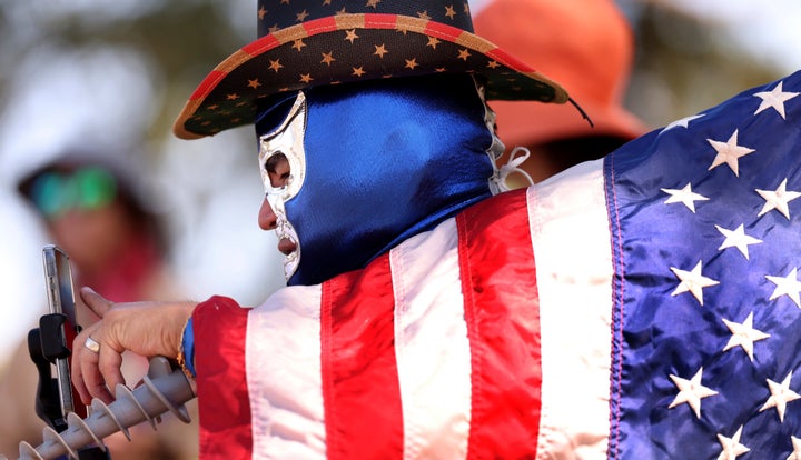 Participante de uma máscara Luchadores, chapéu de cowboy e bandeira dos EUA usa um telefone para fotografar o 