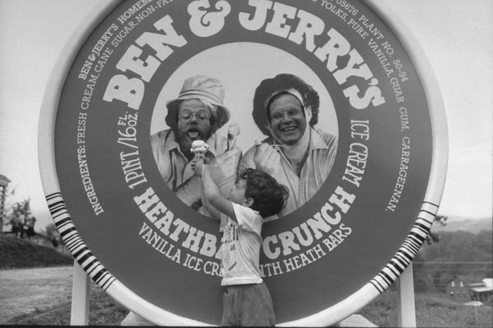 Co-founders Jerry Greenfield (R) & Ben Cohen (L) stick their heads through a sign designed like the top of an ice cream container while boy holds a cone up to them during a company party.