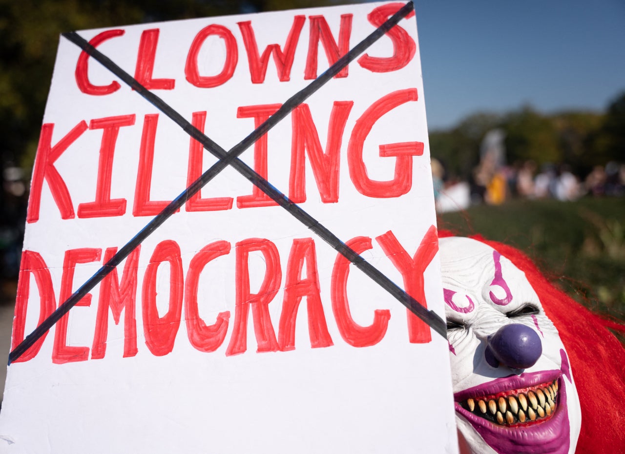 A person in a clown mask holds a sign reading "Clowns Killing Democracy" with an "X" through it near the Tennessee State Capitol building in Nashville, Tennessee.