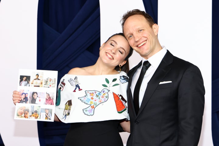 Accurso holds up a photo of some of the kids who contributed drawings to her dress while posing on the red carpet with her husband, Aron.