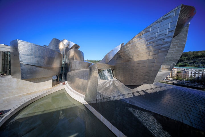 Aerial view of the Guggenheim Museum Bilbao, emphasizing its dramatic, metallic forms that fan out like sculpted wings. The reflective titanium panels capture the sunlight, casting intricate shadows and creating a striking interplay of light across the structure. A shallow pool below mirrors parts of the museum’s architecture, enhancing the sense of fluidity and movement in the design. The vibrant blue sky above contrasts with the museum’s warm metallic tones, highlighting the building’s organic, avant-garde shapes against Bilbao’s landscape.