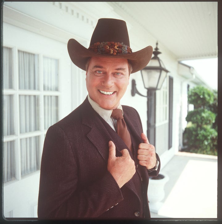 Larry Hagman smiles as he holds up the lapels of his three-piece striped suit, in character as John Ross. 