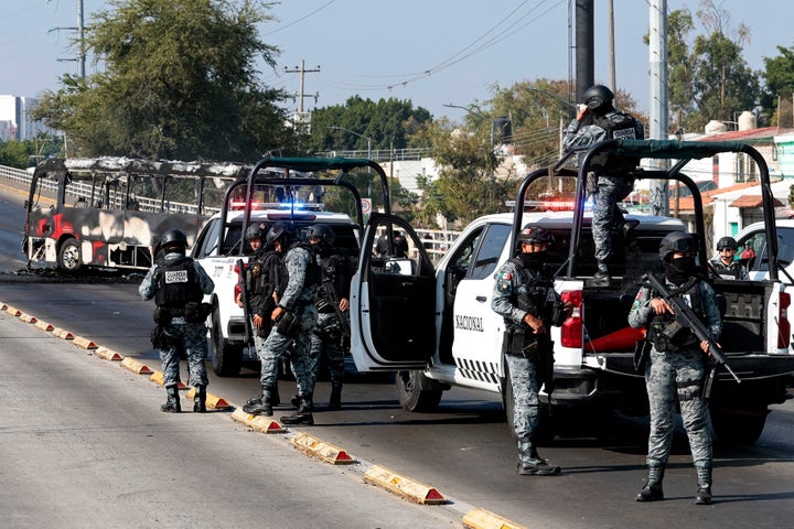 National Guard members stand guard near a burnt bus set on fire by organised crime groups in response to an operation in Jalisco to arrest a high-priority security target, at one of the main avenues in Zapopan, state of Jalisco, Mexico, on February 22, 2026. Armed civilians blocked several roads in the state of Jalisco, in western Mexico, following an operation by federal forces in the town of Tapalpa, local authorities reported. Jalisco, which will host four matches of the upcoming 2026 World Cup, is home to the powerful Jalisco New Generation Cartel (CJNG), and has been rocked by several episodes of violence in recent years. (Photo by Ulises Ruiz / AFP via Getty Images)