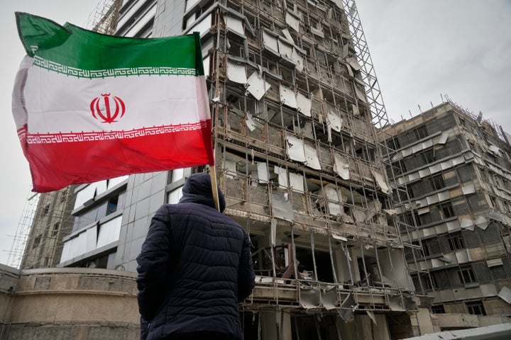 A man holds an Iranian flag as he looks at the damaged façade of Gandhi Hospital, which was hit Sunday when a strike also struck a state TV communications tower and nearby buildings across the street during the ongoing joint U.S.–Israeli military campaign in Tehran, Iran.