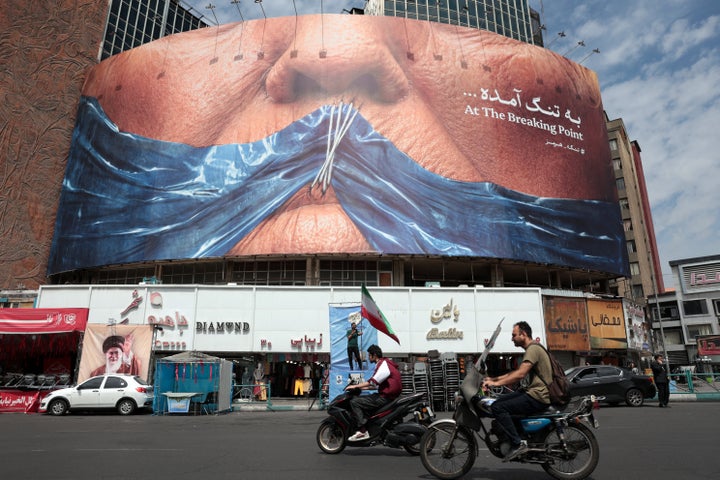 Motorists ride past a billboard in Tehran's Valiasr Square that depicts the Strait of Hormuz being sewn shut over what appears to be President Donald Trump's mouth, on May 2, 2026. Trump said that the United States will begin a program to help stranded ships from