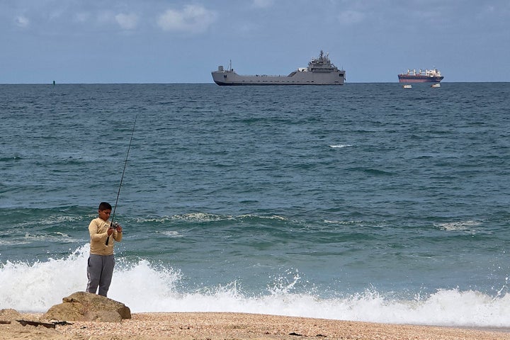 A boy fishes on the shore as an Israeli naval ship (C) brings one of the last Gaza-bound Global Sumud Flotilla vessels toward Ashdod port, about 40 kilometers south of Tel Aviv, on May 20, 2026. Israeli authorities have detained hundreds of activists who set sail from Cyprus last week in an effort to break the Gaza blockade deliver vital humanitarian aid.