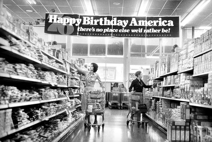 Two women shopping in grocery store under banner reading. "Happy birthday America, there's no place else we'd rather be!" commemorating the US bicentennial.