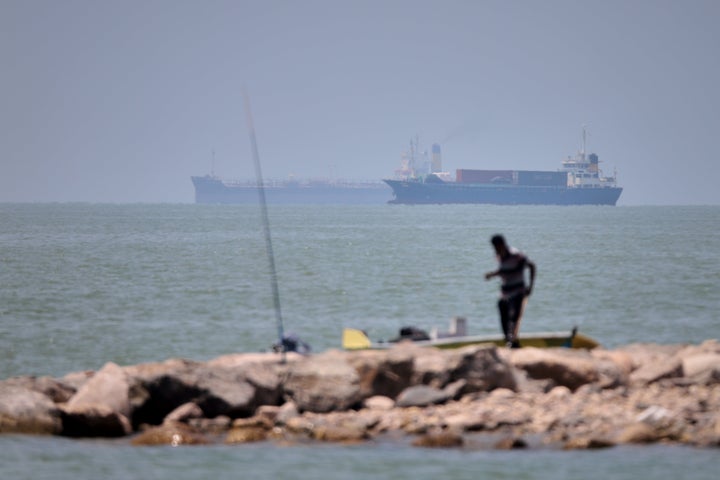 A man stands beside a fishing pole along the shore as cargo ships and commercial vessels are seen in the Strait of Hormuz off Bandar Abbas, Iran, on June 17, 2026.