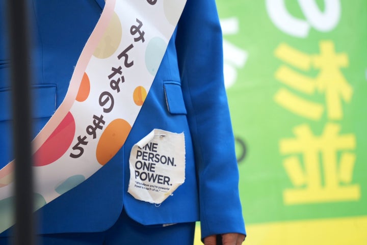「みんなでつくる みんなのまち」のたすきと、「ONE PERSON ONE POWER(ひとりには、ひとり分の力がある)」のワッペンをつけて街宣で話した岸本さん