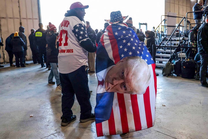 A Trump supporter is draped in a flag emblazoned with the president’s mugshot at a 2024 rally in Waterford Township, Michigan.