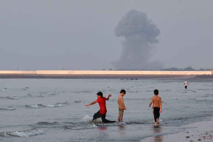 Three children play in the shallow waters of the Strait of Hormuz as smoke rises from a background explosion off the coast of Bandar Abbas, Iran, on July 13, 2026.